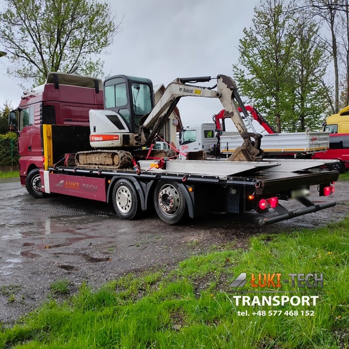 Czerwony samochód ciężarowy przewozi na platformie brudną mini koparkę Bobcat na tle pochmurnego nieba.