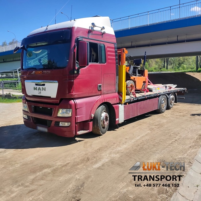 Czerwony ciągnik siodłowy marki MAN z platformą transportową przewożący pomarańczowy wózek widłowy, pod wiaduktem, z logo firmy transportowej w lewym górnym rogu kabiny.