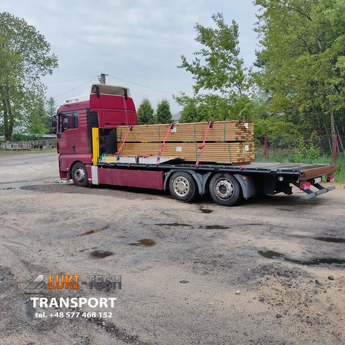 Czerwony samochód ciężarowy z płaską platformą przewożący zabezpieczony ładunek drewna, widoczny na szarym żwirowym podłożu z niewielkimi kałużami i logo firmy transportowej w lewym dolnym rogu.