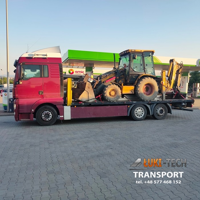 Czerwona ciężarówka z platformą przewozi koparko-ładowarkę na tle stacji benzynowej w Jordanowie. Koparka jest zabezpieczona pasami. Widoczne logo firmy transportowej w prawym dolnym rogu.