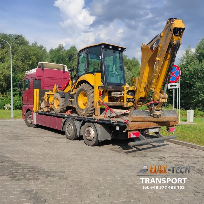 Ciężarówka z naczepą przewożąca żółtą koparko-ładowarkę marki CAT, zabezpieczoną czerwonymi pasami transportowymi, na tle zieleni i błękitnego nieba z chmurami.