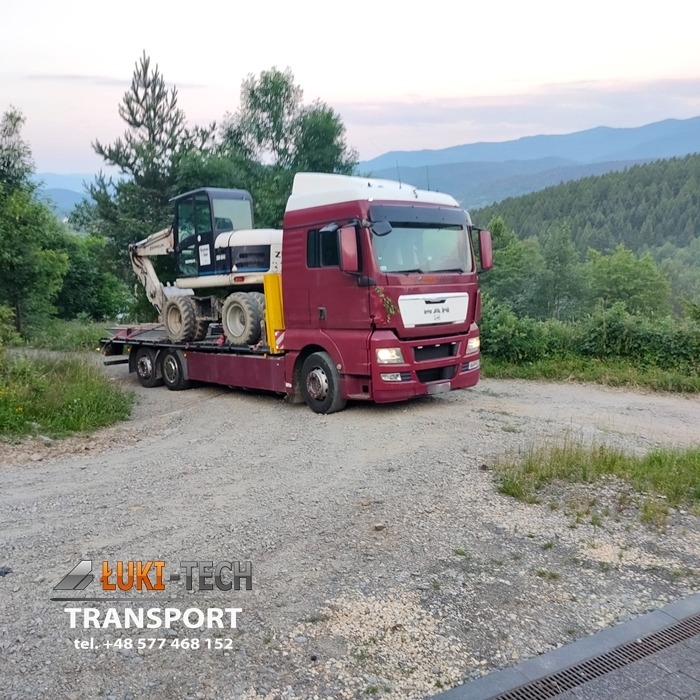 Czerwony ciągnik MAN z naczepą transportuje białą mini koparkę na tle zalesionych wzgórz w pochmurny dzień.