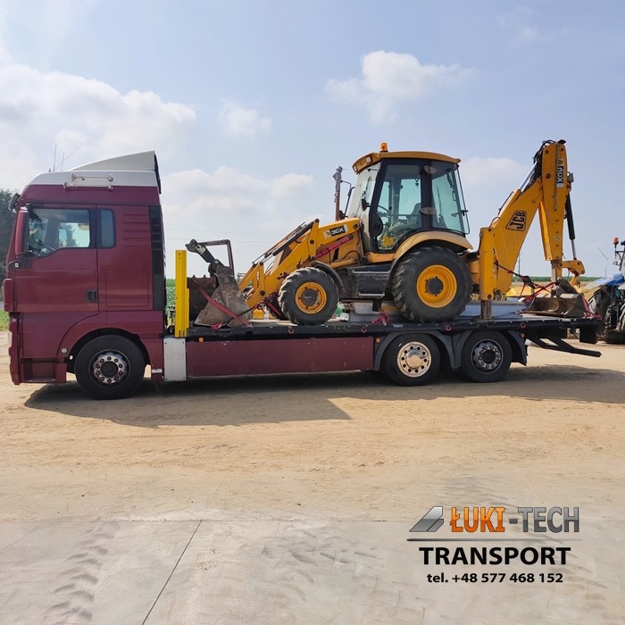 Czerwony samochód ciężarowy z żółtą koparko-ładowarką JCB 3CX na platformie, zabezpieczoną czerwonymi pasami transportowymi, na tle błękitnego nieba.