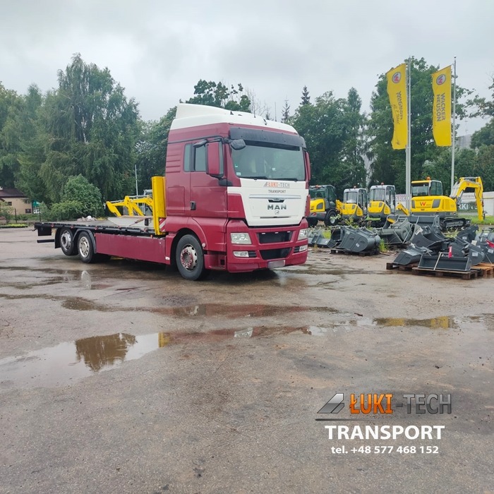 Czerwono-biała ciężarówka MAN z niskopodwoziową platformą, zaparkowana na placu z kałużami, w tle sprzęt budowlany i flagi reklamowe.