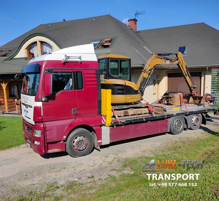 Czerwono-biała ciężarówka MAN z żółtą mini koparką CAT na niskopodwoziowej platformie, zabezpieczona czerwonymi pasami, na tle domu z ciemnym dachem.