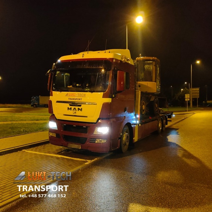 Ciężarówka MAN z logo Luki-Tech Transport przewozi koparkę na niskopodwoziowej platformie transportowej nocą, oświetlona światłami ulicznymi.