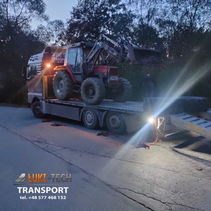 Czerwony traktor z ładowaczem na platformie niskopodwoziowej ciężarówki transportowej, widok z boku, oświetlenie robocze włączone, wieczorny krajobraz w tle.