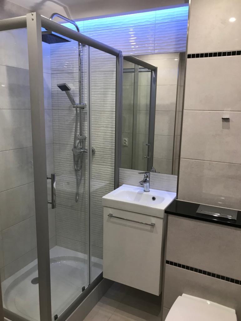 Nowoczesna łazienka z kabiną prysznicową ze srebrnymi profilami, umywalką nablatową na białej szafce, lustrem i podświetleniem LED na suficie.