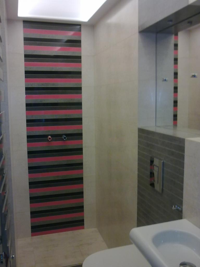 Wykończona łazienka z akcentem w postaci ściany wyłożonej płytkami w poziome pasy: różowe, czarne i szare. Na ścianie nad toaletą szare płytki i wbudowany panel sterujący spłukiwaniem. W lustrze...