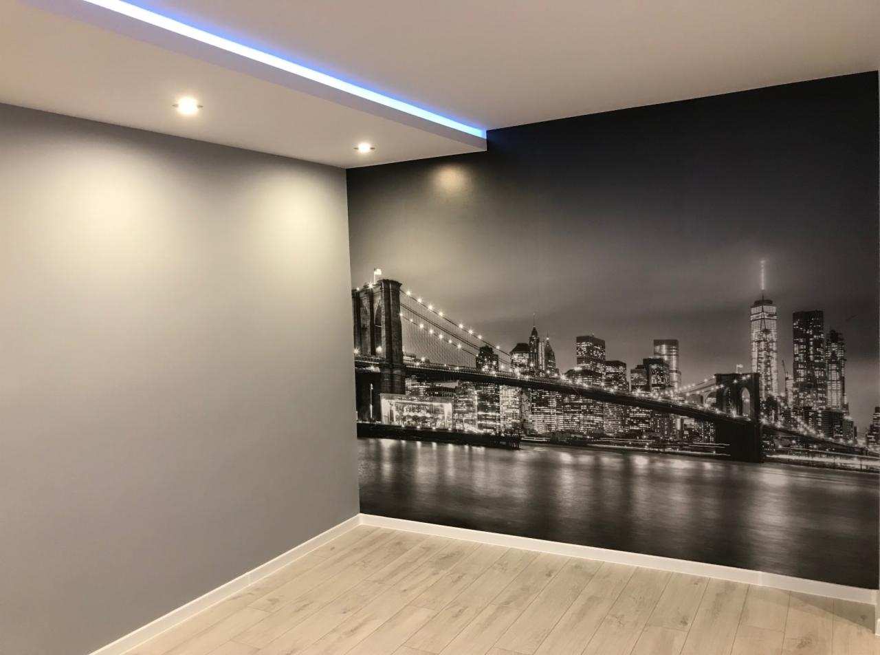 Szara ściana w pokoju z fototapetą przedstawiającą panoramę Nowego Jorku z mostem Brooklińskim, podświetlony sufit z ledami, jasne panele podłogowe.