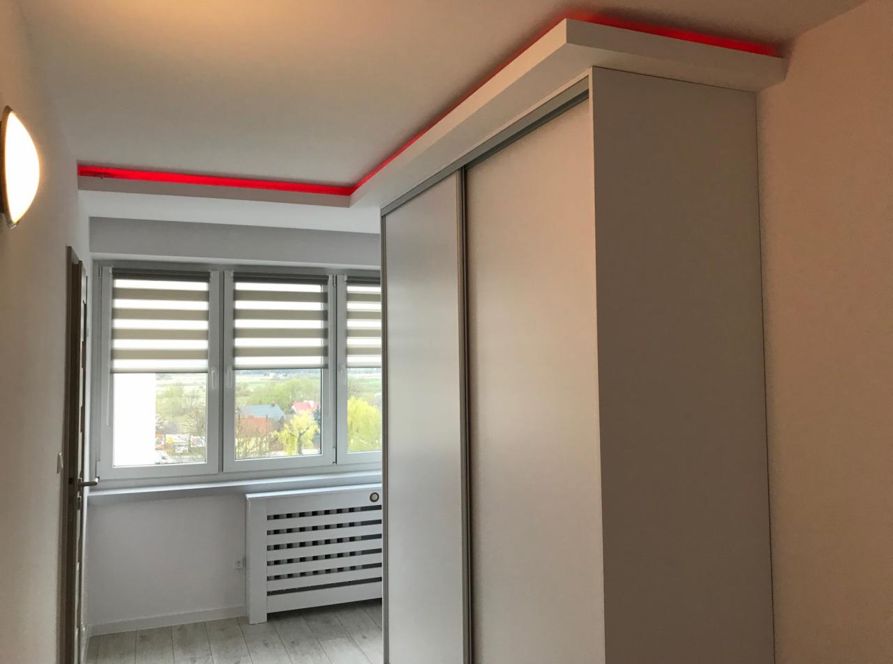 Biała szafa przesuwna z oświetleniem LED w kolorze czerwonym, zamontowana w pokoju z oknem i grzejnikiem.