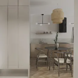 Jasne wnętrze mieszkania z parkietem w jodełkę, widok na kuchnię z okrągłym stołem, krzesła z wiklinowym siedziskiem oraz zabudowa szaf w przedpokoju.