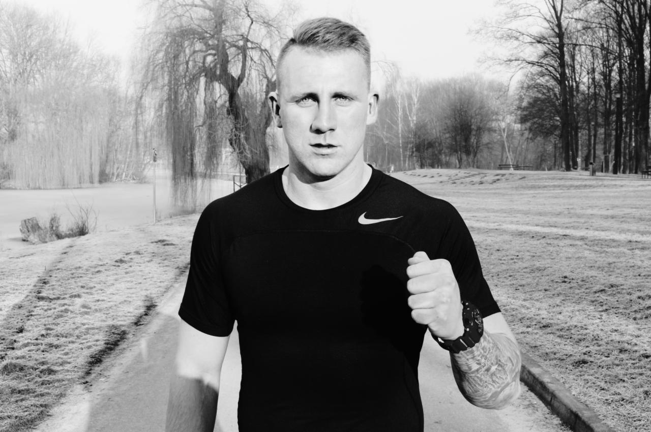 Czarno-białe zdjęcie mężczyzny w sportowej koszulce z logo Nike, stojącego na ścieżce w parku, z widocznym tatuażem na ramieniu i zegarkiem sportowym.