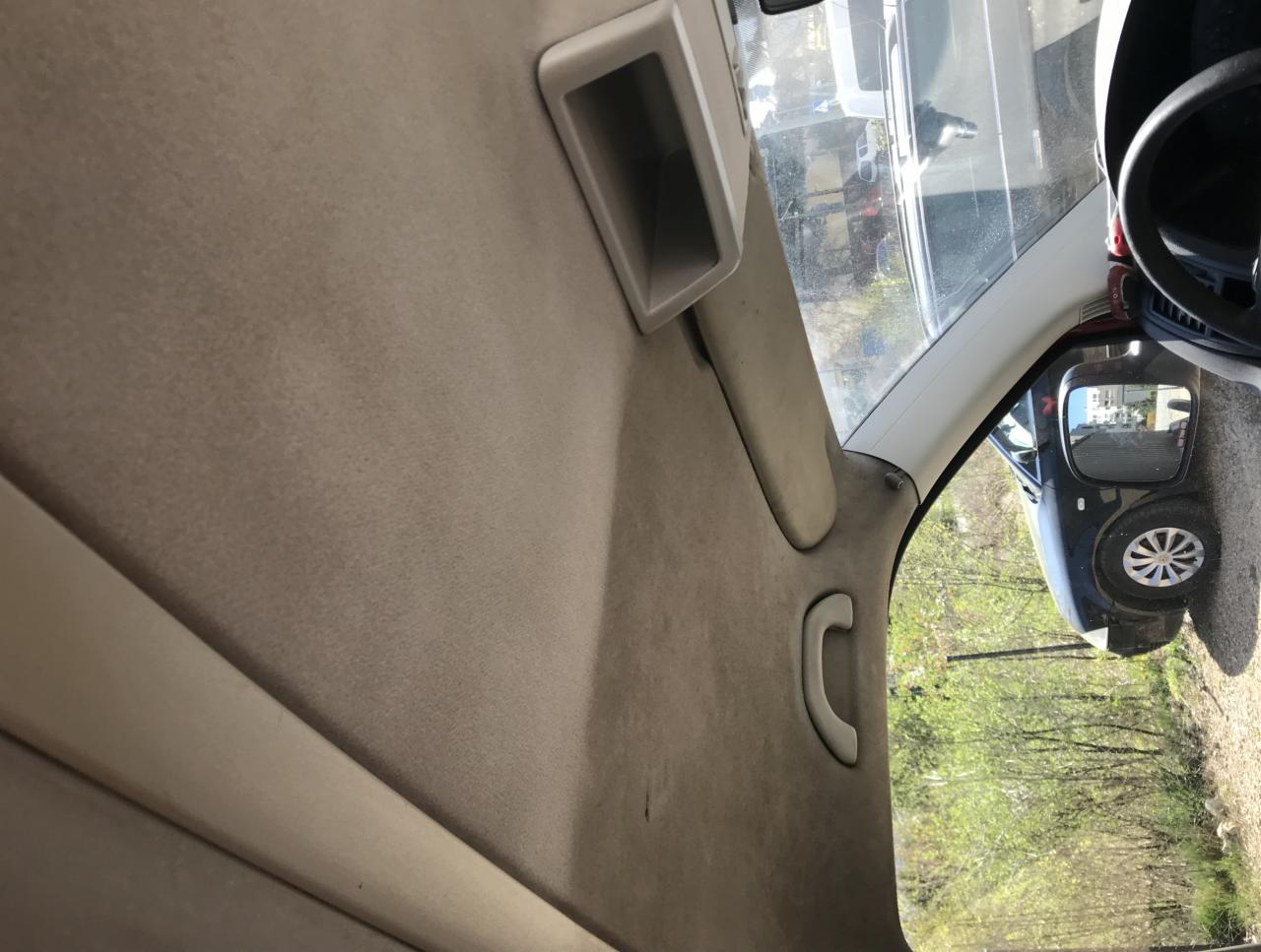 Wnętrze samochodu, widoczna beżowa tapicerka podsufitki z uchwytem, fragment deski rozdzielczej i odbicie samochodu w lusterku bocznym.