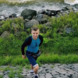 Młody mężczyzna w sportowym stroju, uśmiechnięty, wchodzi po kamiennym zboczu porośniętym trawą, w tle widać fale morskie rozbijające się o kamienie.