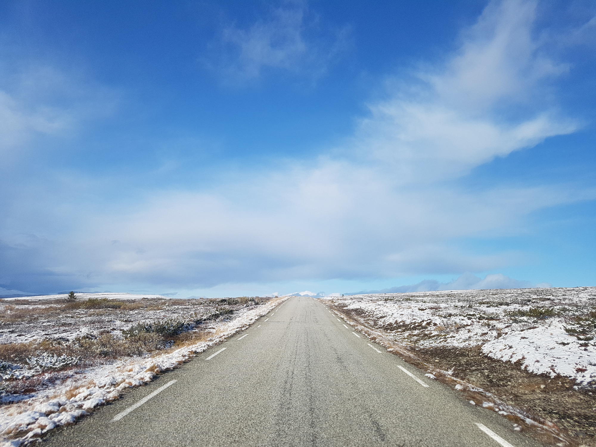 Asfaltowa droga biegnąca przez zimowy krajobraz tundry pod błękitnym niebem z chmurami, z widocznymi śladami opon na nawierzchni.