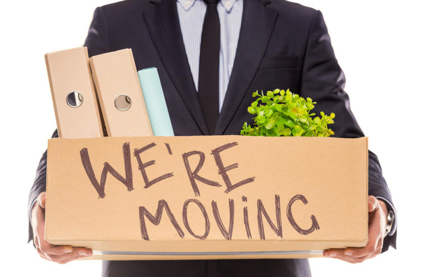 Mężczyzna w garniturze trzyma karton z napisem 'We're moving', zawierający segregatory i roślinę doniczkową.