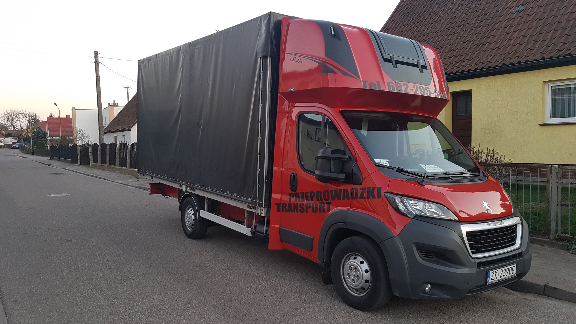Czerwony samochód dostawczy z czarną plandeką, zaparkowany na ulicy w dzielnicy domów jednorodzinnych, z napisem 'Przeprowadzki Transport' i numerem telefonu.