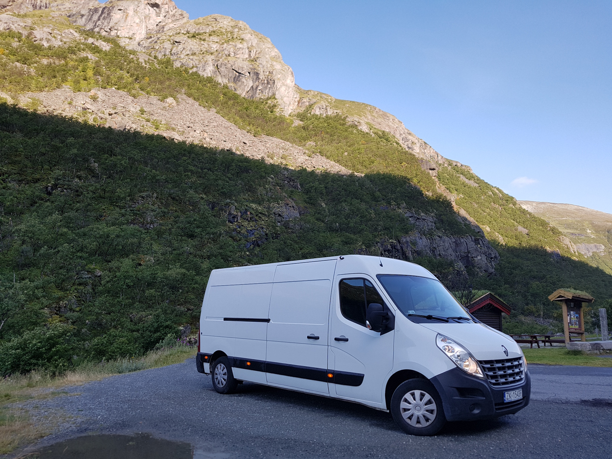 Biały bus Renault Master zaparkowany na tle góry porośniętej zielenią, z widoczną tablicą informacyjną i małą chatką z zielonym dachem w tle.