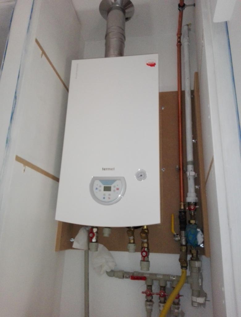 Biały piec gazowy Termet zamontowany na płycie MDF, widoczne rury miedziane i plastikowe, zawory oraz komin odprowadzający spaliny.