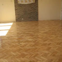 Świeżo położony, lakierowany parkiet w jodełkę, z widocznymi refleksami światła słonecznego w pustym pokoju z ceglaną ścianą akcentującą.