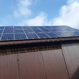 Panel fotowoltaiczny na brązowym dachu budynku gospodarczego z metalowymi wrotami garażowymi, widok z dołu na błękitne niebo z chmurami.