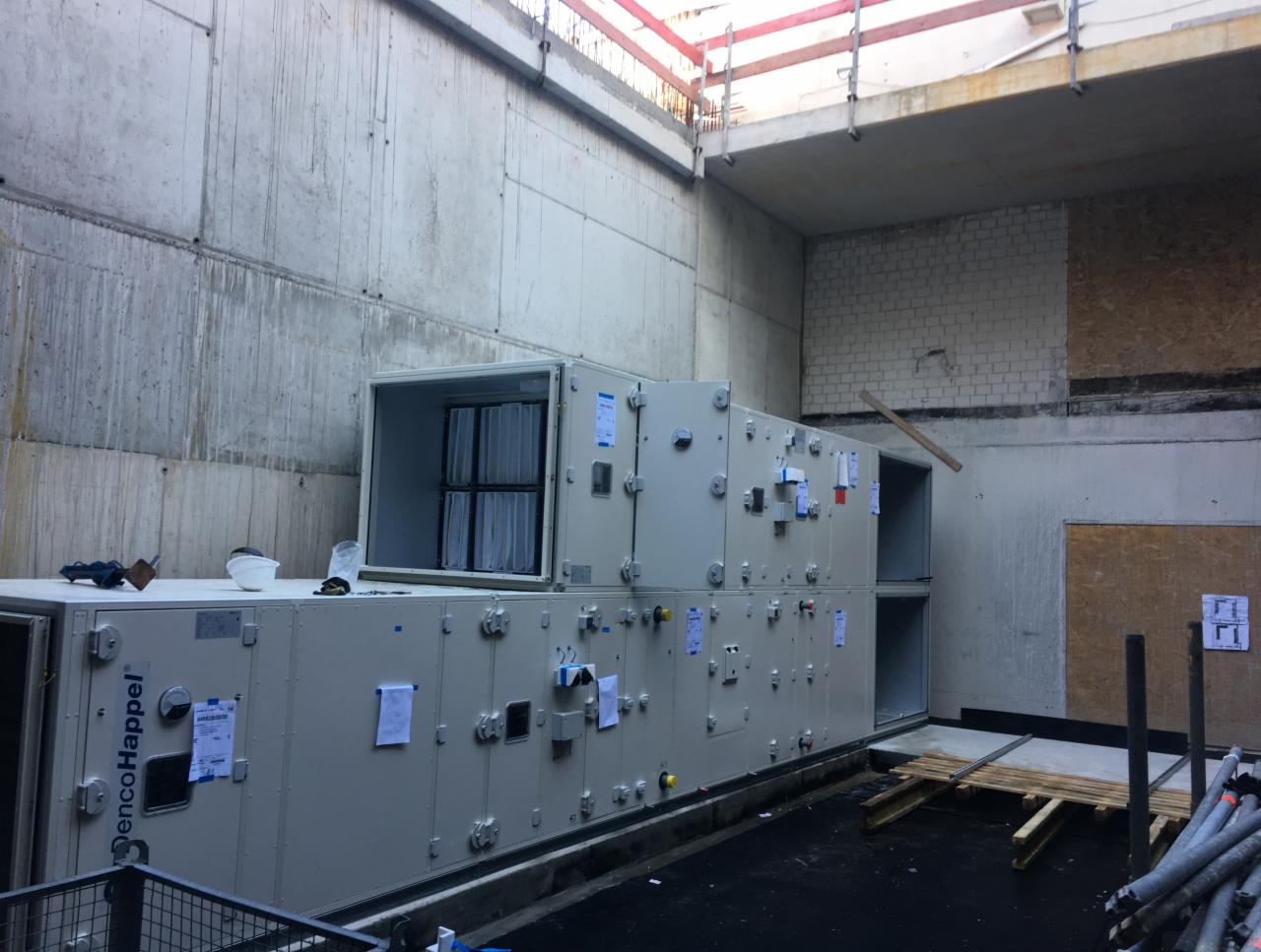Dwa moduły centrali wentylacyjnej DencoHappel ustawione jeden na drugim w surowym, betonowym pomieszczeniu w trakcie montażu. Widoczne filtry w otwartym module, elementy montażowe i notatki...