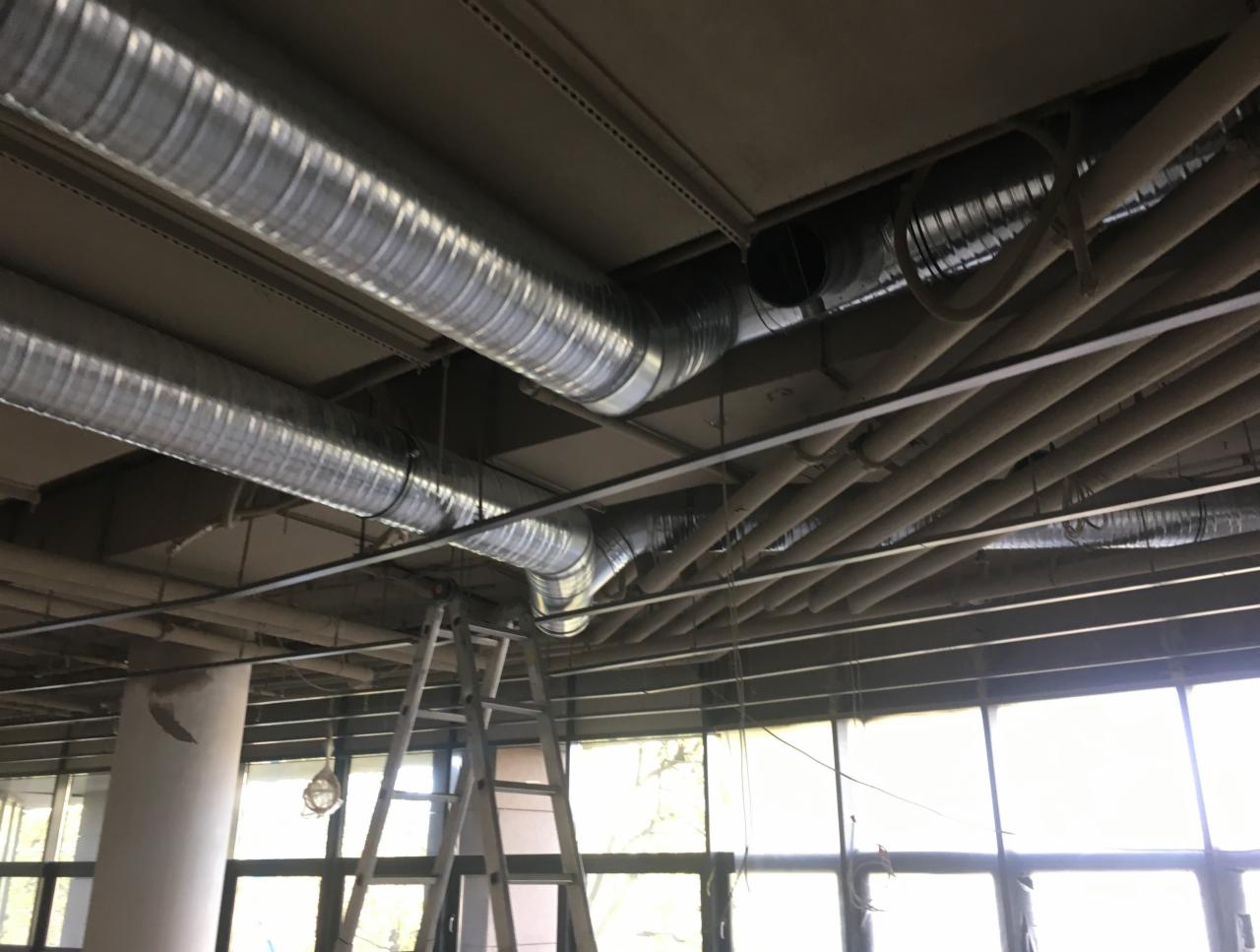 Instalacja wentylacyjna z rurami spiro pod stropem budynku komercyjnego, widoczna drabina aluminiowa i okna.
