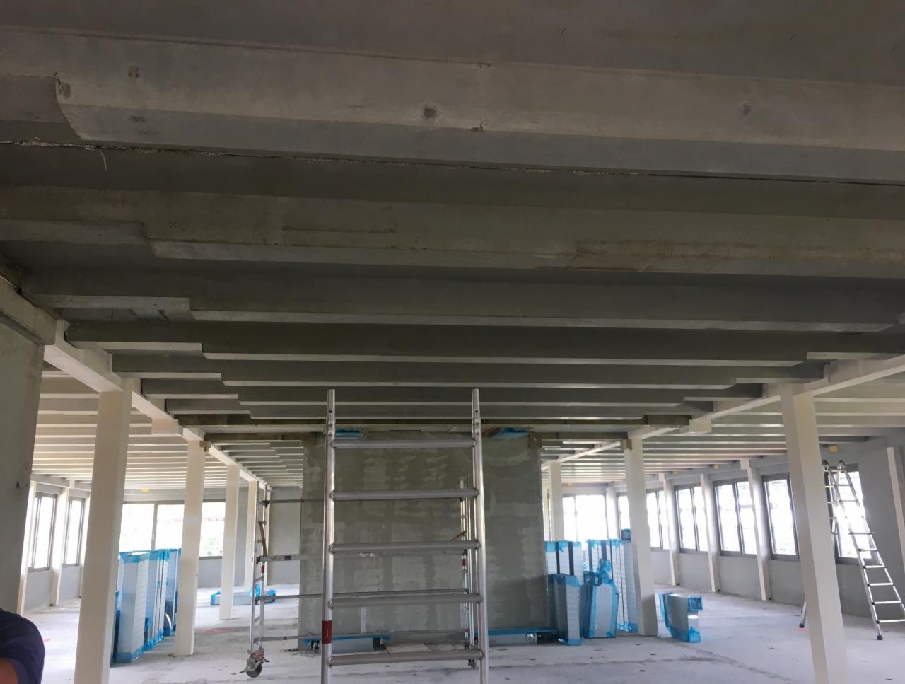 Wnętrze budynku komercyjnego w trakcie montażu instalacji wentylacyjnej, widoczne betonowe stropy, białe kolumny nośne, okna oraz drabina montażowa. Część elementów zabezpieczona folią ochronną.