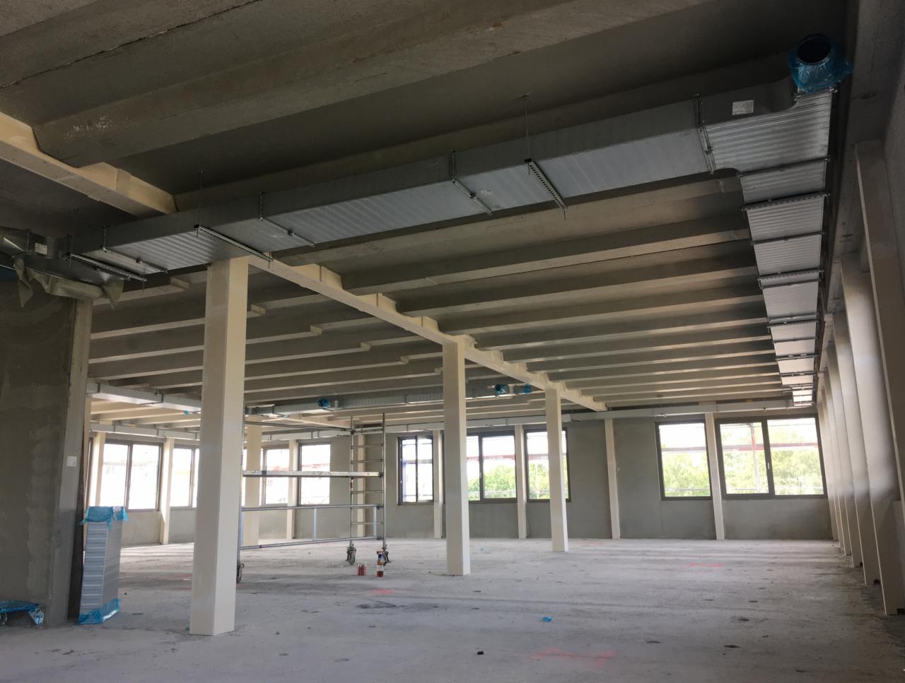 Wnętrze budynku komercyjnego w trakcie instalacji systemu kanałów wentylacyjnych pod sufitem, widoczne kolumny nośne i okna.