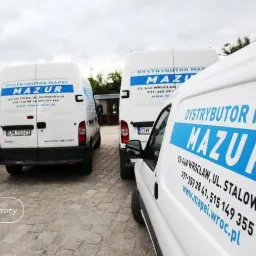 Flota białych samochodów dostawczych z niebieskim logo dystrybutora MAPEI Mazur, zaparkowanych na brukowanym placu pod pochmurnym niebem.