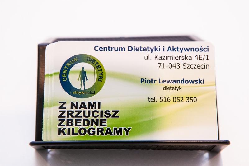 Wizytówki Centrum Dietetyki i Aktywności w czarnym, plastikowym podajniku, z hasłem 'Z Nami Zrzucisz Zbedne Kilogramy' na pierwszym planie.