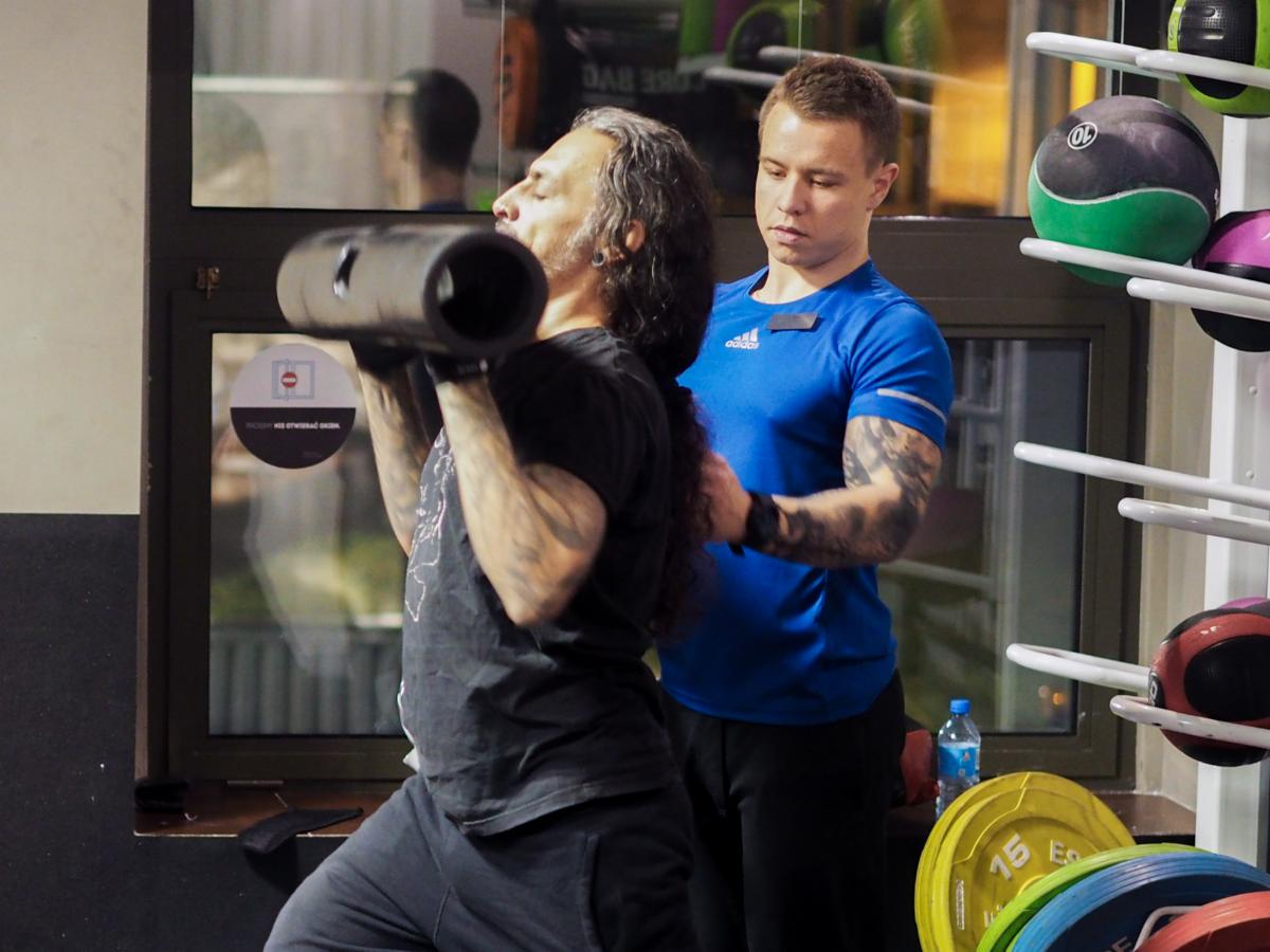 Mężczyzna z długimi włosami ćwiczy z obciążeniem w kształcie walca pod nadzorem trenera personalnego w niebieskiej koszulce w siłowni.