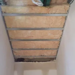 Izolacja z wełny mineralnej zamontowana w metalowym stelażu poddasza, widoczne przewody elektryczne i fragment ściany pomalowanej na biało.