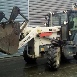 Biało-szara koparko-ładowarka Terex TLB890 z brudną łyżką z przodu, stojąca na mokrym placu przed budynkiem z blachy falistej.