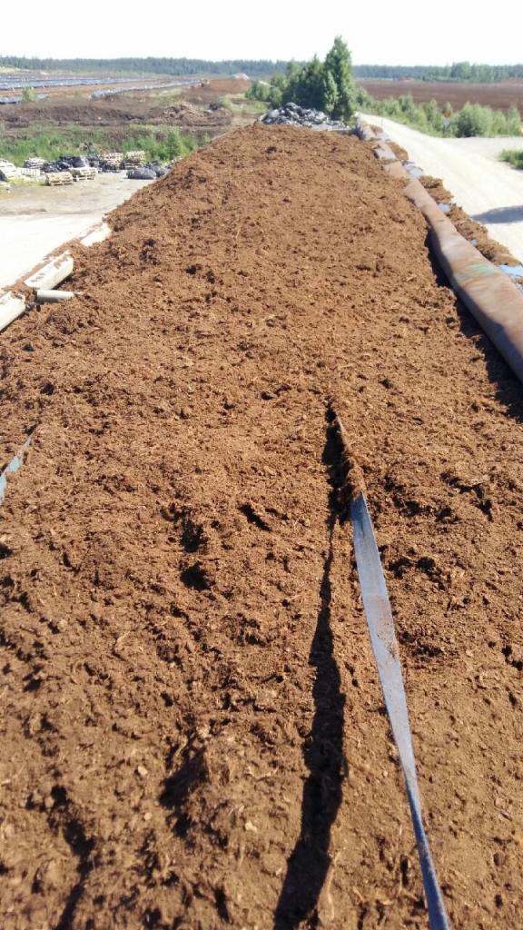 Widok z góry na przyczepę wypełnioną brązowym, sypkim materiałem przypominającym torf lub kompost, z narzędziem wbitym w jego powierzchnię, w tle widoczne pole i drzewa.