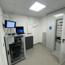 Serwerownia z szafą rack, monitorem, laptopem i otwartą szafą elektryczną w pomieszczeniu z białymi ścianami i szarą podłogą.