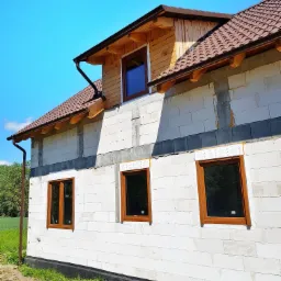 Surowy stan elewacji domu jednorodzinnego z zamontowanymi oknami o brązowych ramach, widoczne bloczki z betonu komórkowego i ocieplenie fundamentów.