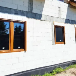 Montaż okien z brązowymi ramami w surowej ścianie z białych bloczków, widoczne zabezpieczenia montażowe i fragmenty konstrukcji dachu.