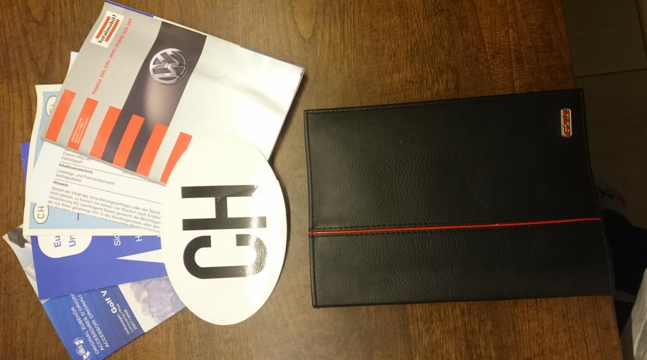Dokumenty Volkswagen, w tym instrukcje, broszury i naklejka CH na drewnianym stole obok czarnego etui z czerwoną linią i logo GTI.