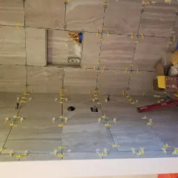 Ściany łazienki w trakcie układania beżowych płytek imitujących marmur, widoczne krzyżyki dystansowe, poziomica i wnęka wykończona płytkami z logo 'TRULY'.