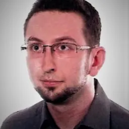 Portret mężczyzny w okularach i ciemnej koszuli na jasnym tle, z krótką brodą i wąsami.