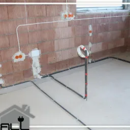 Surowa ściana z cegły z zamontowanymi puszkami elektrycznymi i przewodami, częściowo zamurowane, widoczne przewody pod podłogą i logo firmy w rogu.