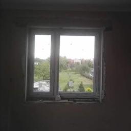 Nasz dom - Nowe, białe okno PCV z podwójnymi szybami, zamontowane w surowym otworze ściennym, z widokiem na zielony ogród i zabudowania w tle.