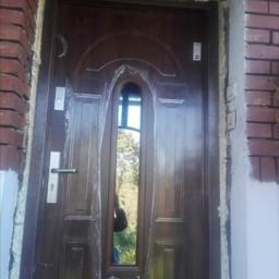 Nasz dom - Brązowe drzwi wejściowe z szybą, zabezpieczone folią, osadzone w murze z widoczną pianką montażową i świeżym malowaniem ściany.