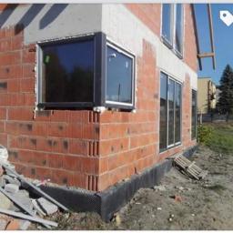 Nasz dom - Widok z zewnątrz na budynek w trakcie montażu okien z czarnymi ramami, widoczny mur z czerwonej cegły, czarna izolacja fundamentu i częściowo zagospodarowany teren.