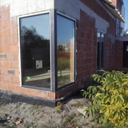 Nasz dom - Nowe, duże okno narożne z czarną ramą w surowym stanie budynku z cegły, widoczne czarne zabezpieczenie fundamentów i ziemia.