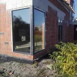 Nowe, duże okno narożne z czarną ramą w surowym stanie budynku z cegły, widoczne czarne zabezpieczenie fundamentów i ziemia.