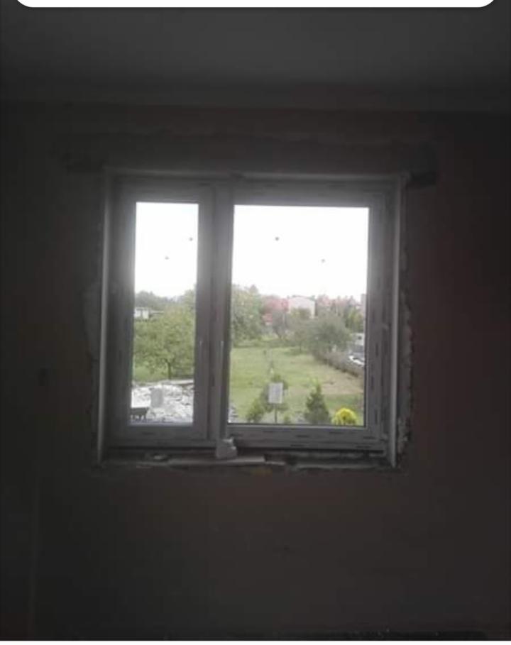 Nowe, białe okno PCV z podwójnymi szybami, zamontowane w surowym otworze ściennym, z widokiem na zielony ogród i zabudowania w tle.