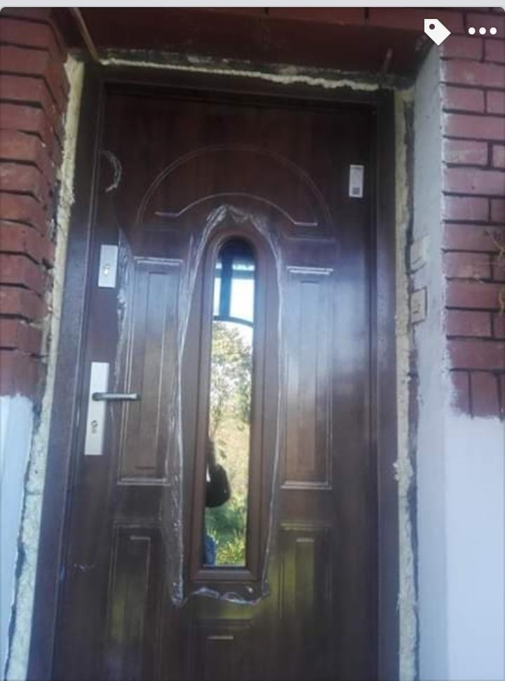 Brązowe drzwi wejściowe z szybą, zabezpieczone folią, osadzone w murze z widoczną pianką montażową i świeżym malowaniem ściany.